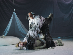 Aktor w kostiumie kudłatego biało-szarego psa stoi nad leżącą aktorką w zielonym kostiumie węża i ją dusi. Wąż łapie powietrze przez szeroko otwarte usta.