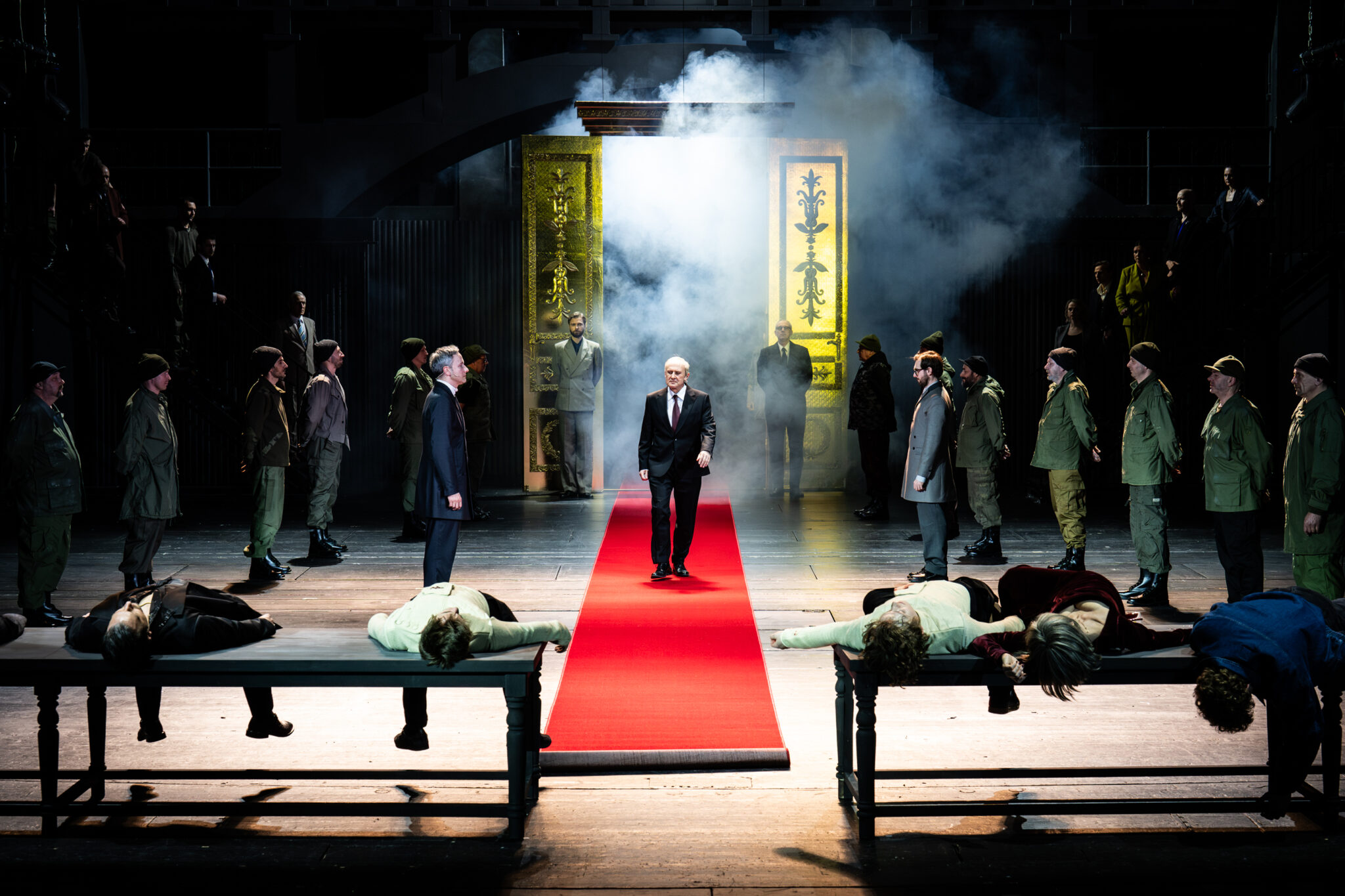 Scena zbiorowa. Na środku czerwony dywan, w głębi złote wrota. Po dywanie idzie mężczyzna w czarnym garniturze. Na proscenium stoją dwie ławki - na nich leżą ciała.