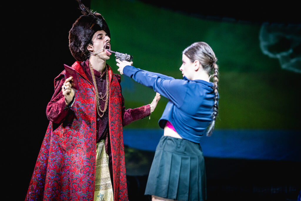 Dwie postaci stojące blisko siebie. Z lewej strony widzimy mężczyznę w staropolskim kontuszu. Z prawej dziewczynę w niebieskiej bluzie, która celuje do niego z pistoletu.