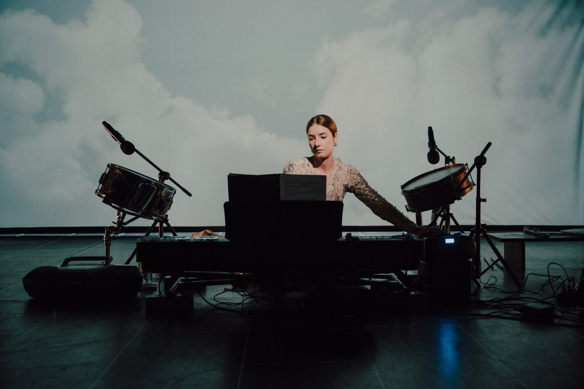 Kobieta siedzi na podłodze za elektrycznym pianinem. Po bokach stoją bębny na statywach. W tle tkanina z wyświetlanymi na niej chmurami.