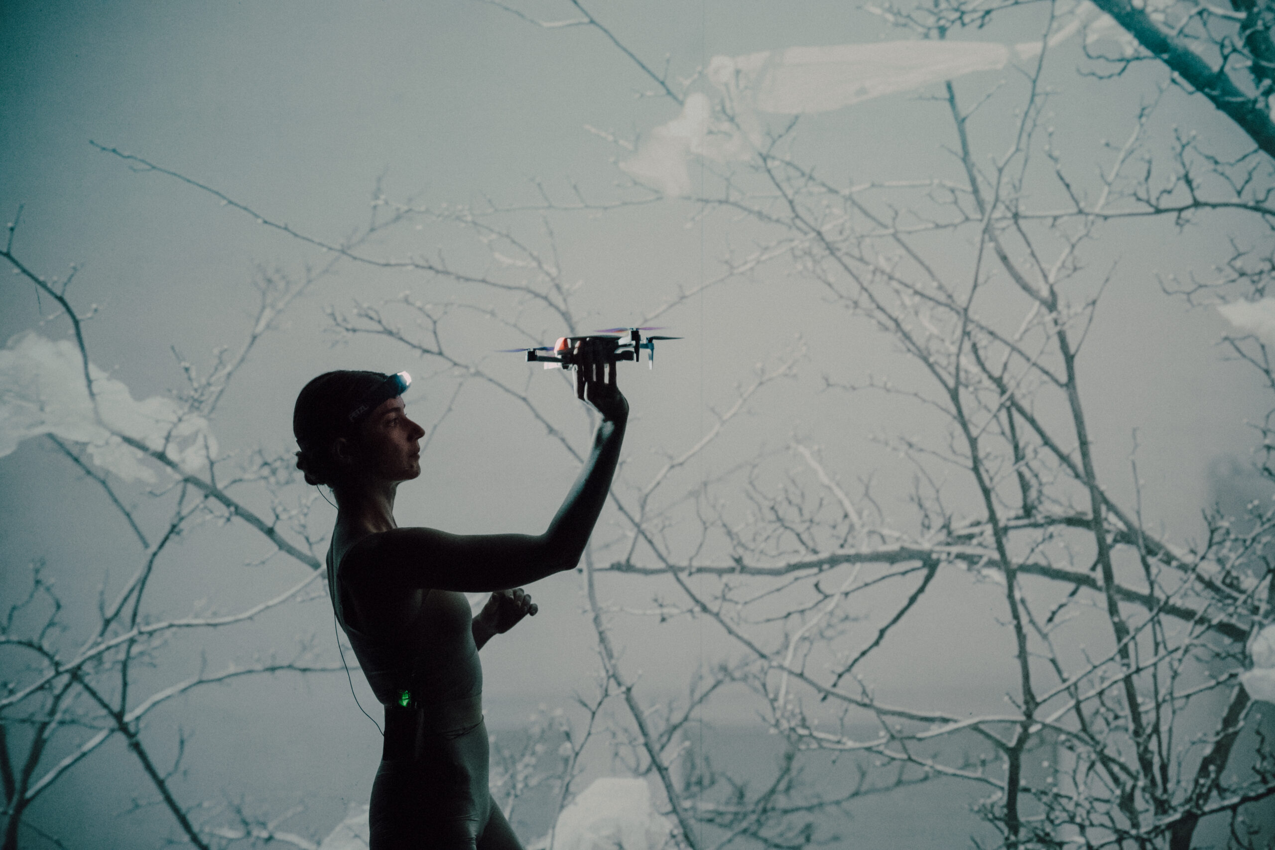 Aktorka stoi trzymając w wyciągniętej ręce drona. W tle ekran z wyświetlanym obrazem bezlistnych gałęzi.