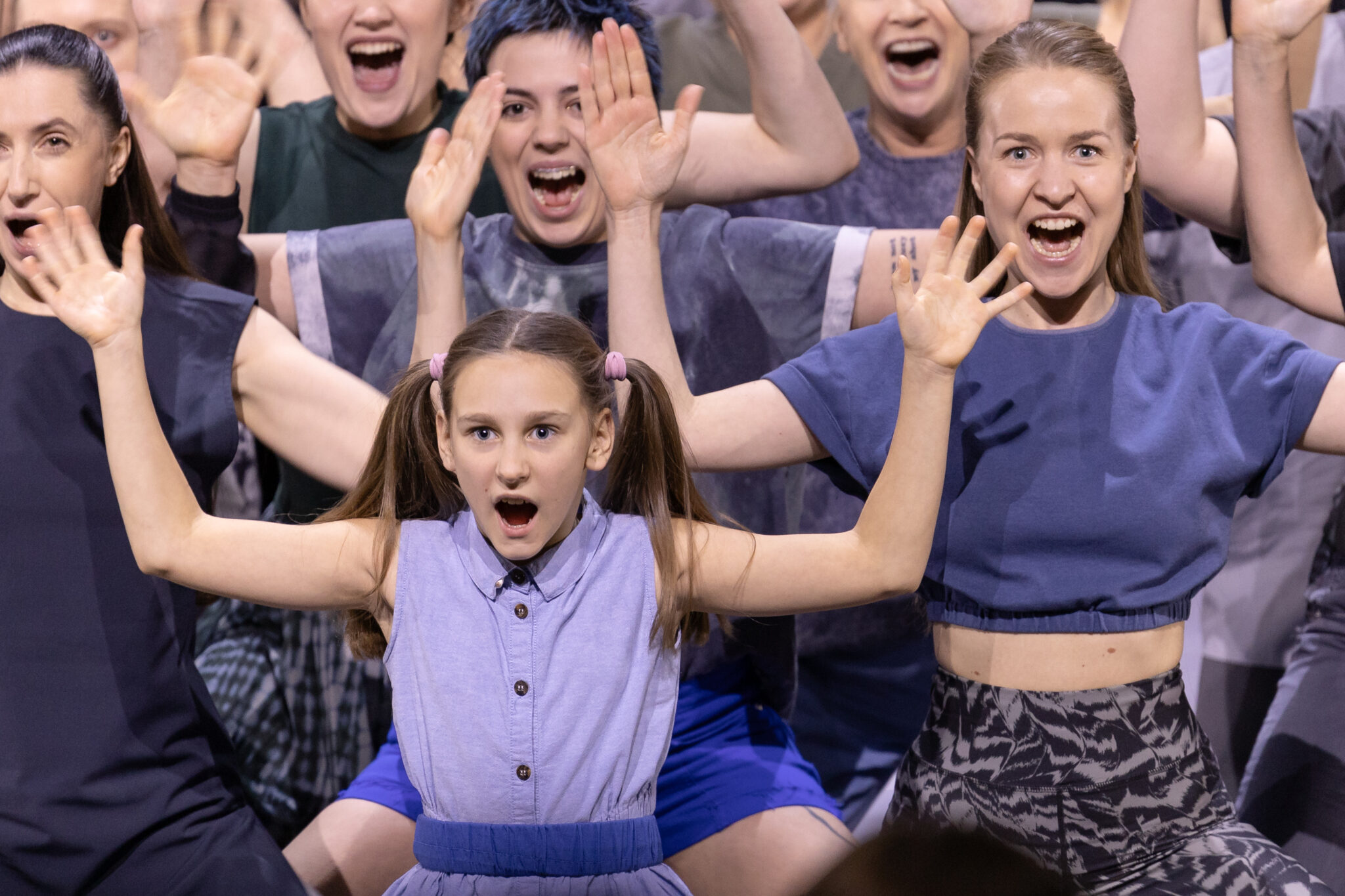 Scena zbiorowa widoczna frontalnie - grupa kobiet śpiewa z uniesionymi rękoma. Na pierwszym planie kilkuletnia dziewczynka w niebieskiej sukience z dwiema kitkami.