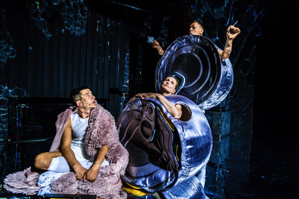 Troje postaci z czego dwie w kostiumach przypominających konserwowe puszki. Aktor na pierwszym planie siedzi po turecku.