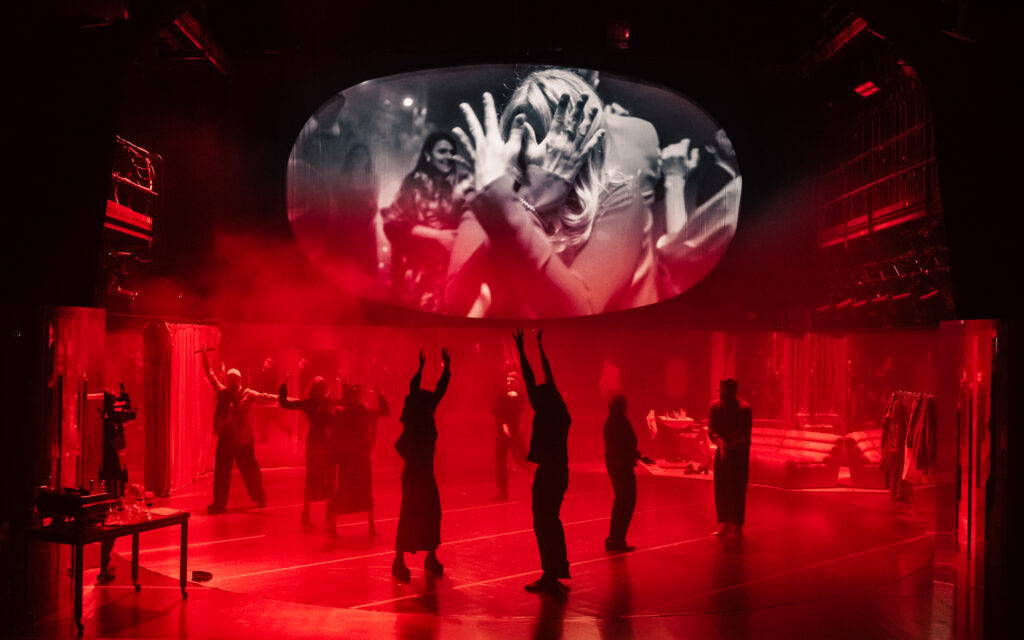 Na górnym planie ekran, na którym widać kobietę zasłaniającą twarz rękoma. Na dolnym planie ciemne sylwetki siedmioro aktorów z uniesionymi do góry rękoma.