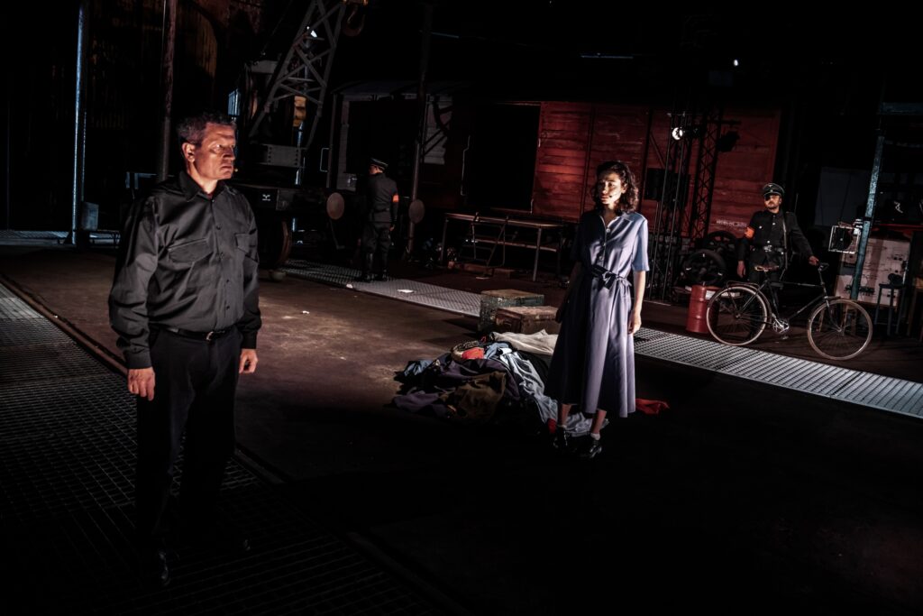 Mężczyzna i kobieta stoją na ciemnej scenie. Jedna osoba leży.