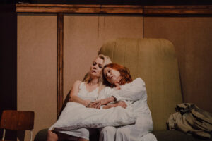 W centrum kadru siedzą dwie kobiety: starsza i młodsza, obydwie w białych koszulach nocnych. Przytulone do siebie.