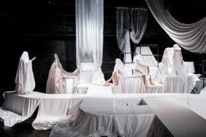 Na scenie widać skomplikowany układ podestów przykrytych białym płótnem. W niektórych miejscach siedzi lub stoi sześcioro aktorów w białych kostiumach duchów.