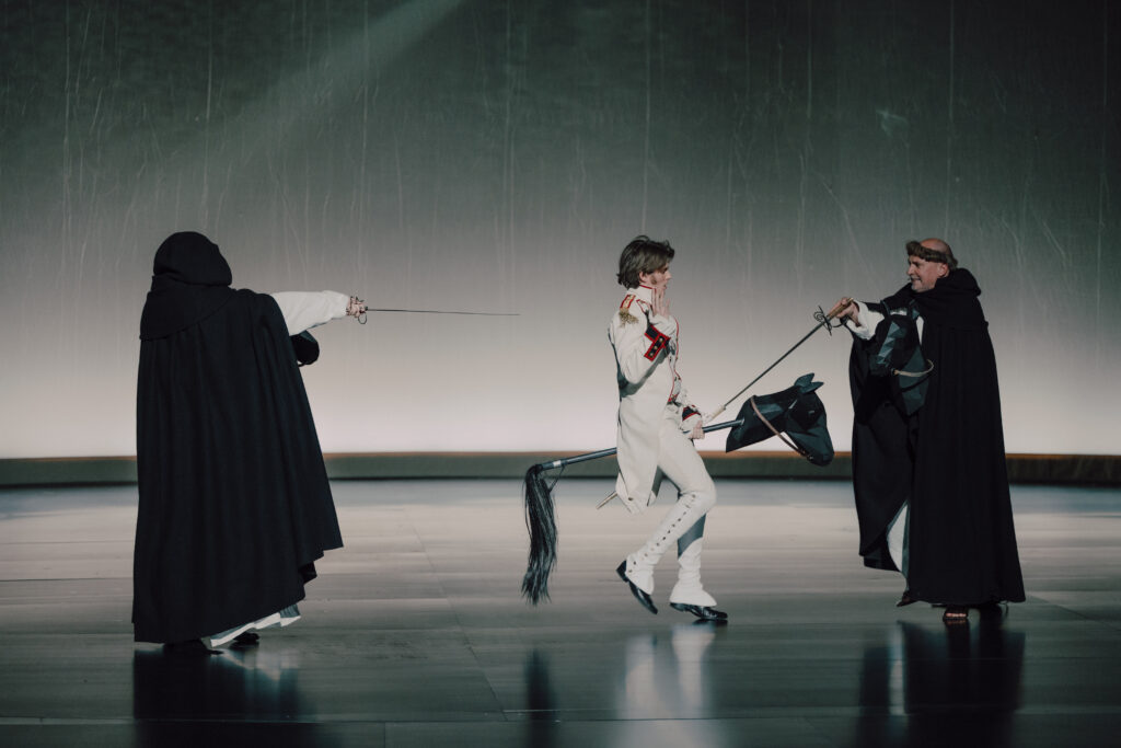 Widzimy trzy postaci. W centrum ubranego w biały mundur aktora, który trzyma między nogami kij symbolizujący konia. Przed sobą i za sobą ma zakonników w czarno-białych strojach. Mierzą w niego szpadami.