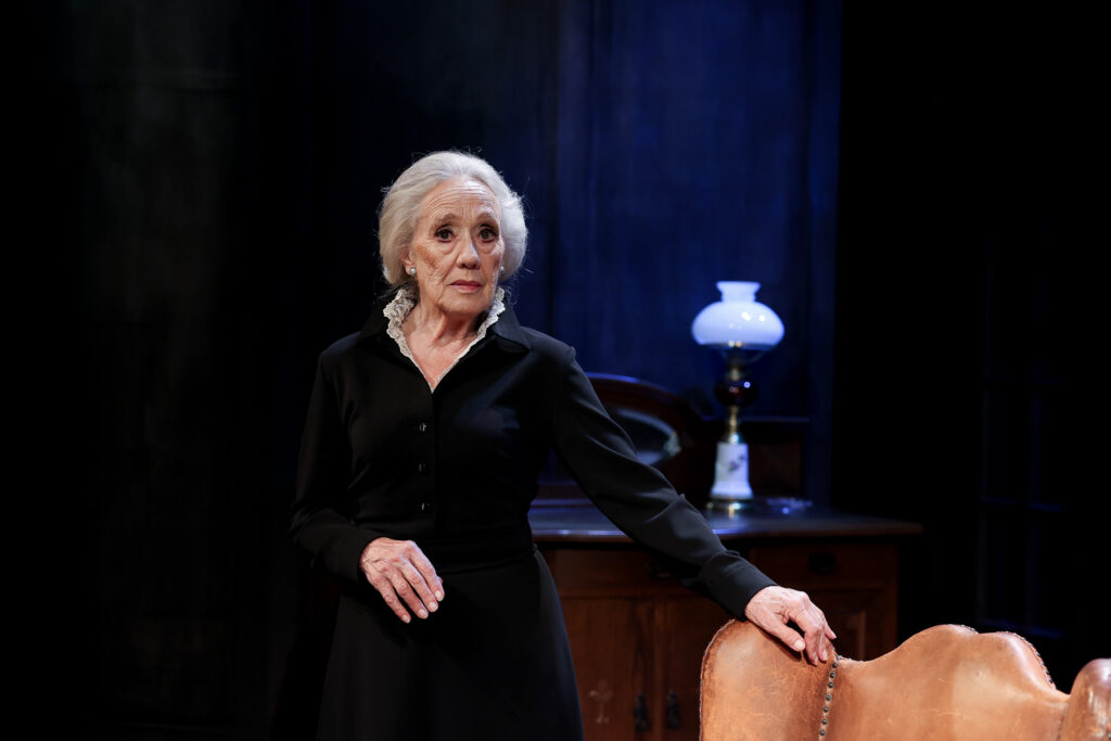 W centrum widać aktorkę w czarnym stroju, która patrzy w obiektyw, trzymając rękę na oparciu fotela.