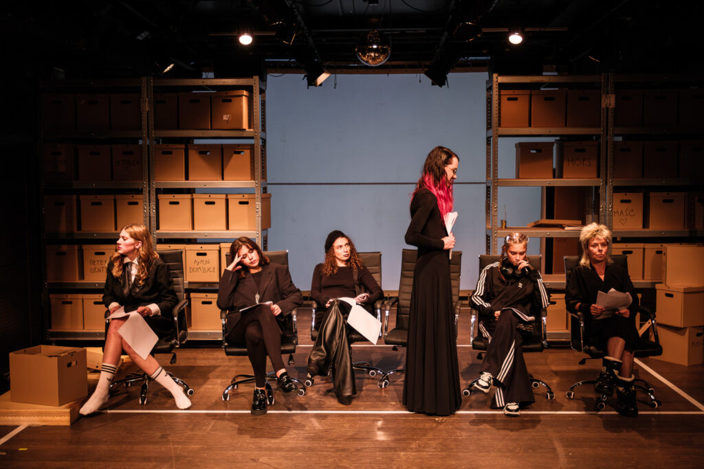 W tle sceny widać magazynowe regały z kartonowymi pudłami. Przed nimi na ustawionych w rzędzie biurowych fotelach siedzi pięcioro aktorek w ciemnych strojach. Szósta stoi profilem do obiektywu.