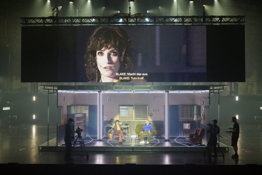 Na zdjęciu widać szerokie ujęcie sceny. W jej centrum znajduje się dwupoziomowa scenografia. Jej górną część stanowi ekran, na którym wyświetlane jest zbliżenie na aktorkę siedzącą w dolnej części (wraz z drugim aktorem).