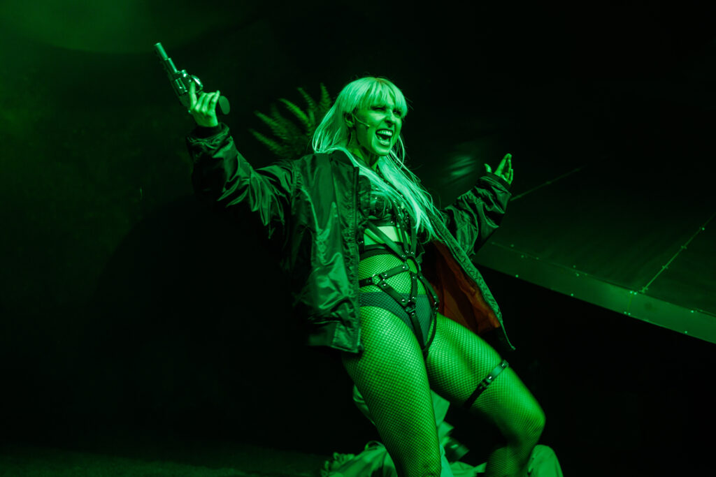 Scena zalana jest zielonym światłem. W centrum zdjęcia widać aktorkę z uniesionymi rękoma. W prawej dłoni trzyma pistolet.