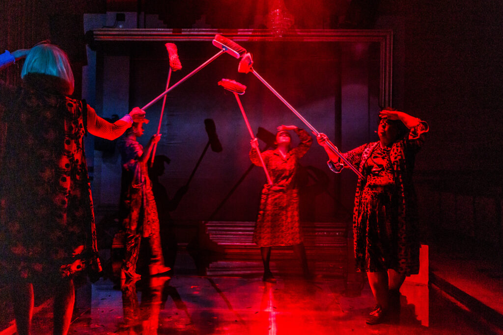 Na zdjęciu widać czworo postaci z uniesionymi do góry szczotkami do zamiatania. Scenę spowija czerwone światło.