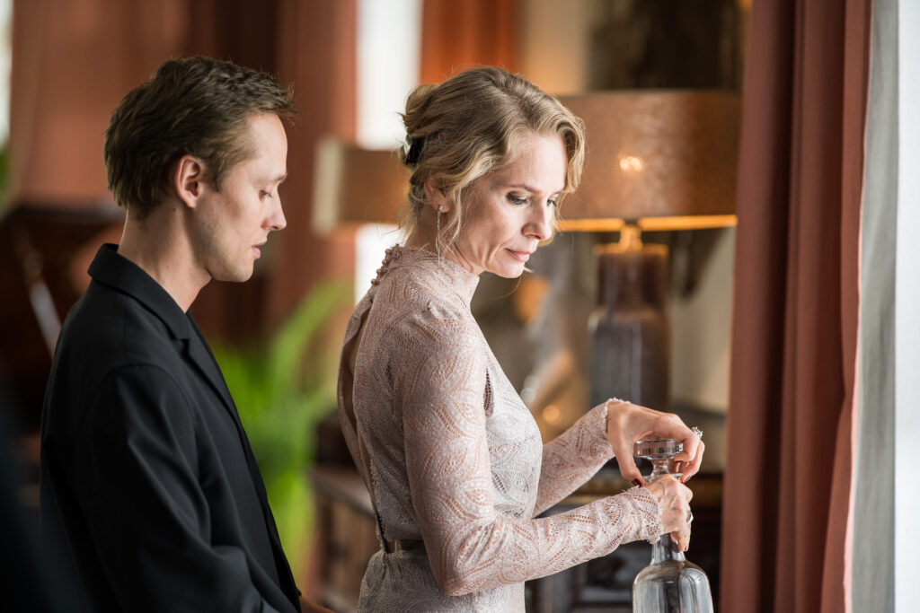 W centrum zdjęcia widzimy aktorkę i aktora, zwróconych profilem. Mężczyzna patrzy na plecy kobiety.